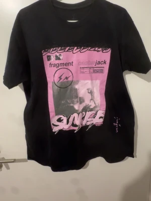 Cactus Jack Travis Scott merch - Cool svart t-shirt med stort rosa grafiskt tryck på framsidan där det står 'fragment' och 'cactus jack'. T-shirten har rund hals och korta ärmar. Perfekt för dig som gillar streetwear och unika prints.