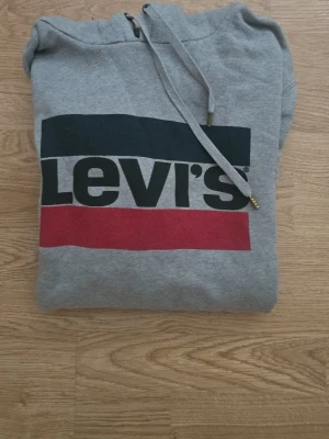 Grå Levi's hoodie med logga - Säljer en grå hoodie från Levi's med stor logga i svart, vitt och rött på bröstet. Hoodien har huva med dragsko och magficka.