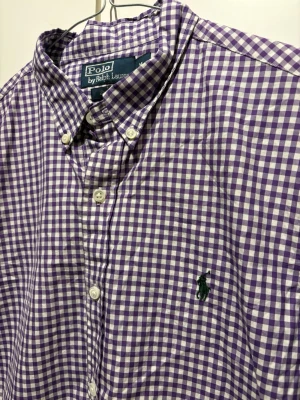Lila rutig skjorta från Polo Ralph Lauren - Säljer en klassisk lila och vit rutig skjorta från Polo Ralph Lauren med button-down krage och broderad logga på bröstet. Skjortan har långa ärmar och vita knappar framtill. Perfekt för en stilren och avslappnad look.