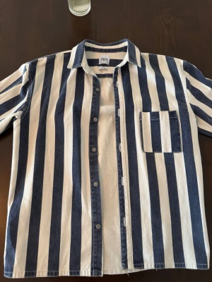 Jeans jacka Zara overshirt  - Snygg overshirt från ZARA med breda vertikala ränder i marinblått och vitt. Skjortan har en klassisk krage, knappstängning framtill och en bröstficka. Tillverkad i ett kraftigare bomullstyg som ger en avslappnad men trendig look.