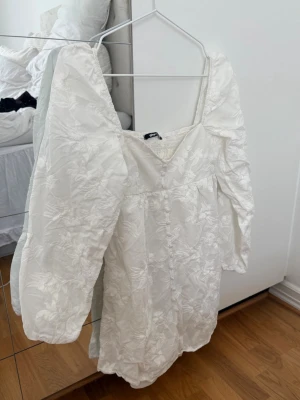 Vit kort klänning med puffärm - Säljer en vit kort klänning med puffiga långärmar och v-ringad hals. Klänningen har ett subtilt blommigt mönster i tyget och en romantisk känsla. Perfekt för sommarens alla tillfällen och har en luftig passform.