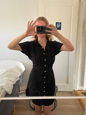 Svart skjortklänning med knappar - En svart skjortklänning med korta ärmar och vita knappar längs hela framsidan. Klänningen har klassisk krage och är tillverkad i ett lätt tyg som passar perfekt till varma dagar. Enkel och stilren design som är lätt att matcha.