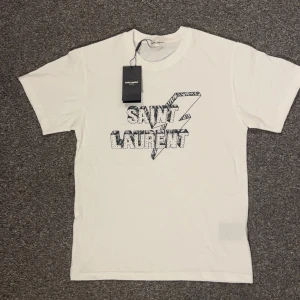 Vit Saint Laurent t-shirt med tryck - Vit t-shirt från Saint Laurent med svart grafiskt tryck och logga på bröstet. Slim passform. Storlek S men skulle säga att den passar xs också. 63 cm lång, 43 cm bred. ‼️ Lapparna kommer tyvärr inte med‼️