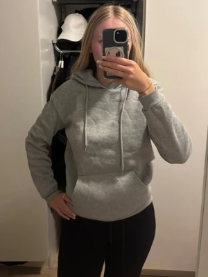 Ljusgrå hoodie med huva - Säljer en enkel och stilren ljusgrå hoodie med huva och dragsnören. Hoodien har en klassisk känguruficka framtill och ribbade muddar vid ärmslut och nederkant. Perfekt för en avslappnad och bekväm stil. Den är tunn så skönt att använda till höst/vår. 