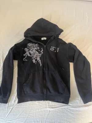Bvaldi hoodie - Säljer en svart hoodie med dragkedja och huva från bvaldi. På bröstet finns ett stort motiv av en ängel i strasstenar samt textdetalj på ena sidan. Hoodien har fickor framtill och justerbara snören i huvan. Perfekt för dig som gillar streetwear och unika detaljer.