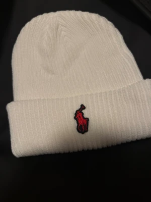 Vit  mössa Polo Ralph Lauren - Vit ribbstickad mössa från Polo Ralph Lauren med uppvikt kant och klassisk röd logga framtill. Tillverkad i mjukt material som håller dig varm och stilsäker under kalla dagar. Enkel och clean design som passar till det mesta.