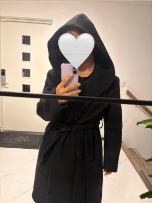Svart kappa med luva  - Stilren svart kappa med luva och knytskärp i midjan. Kappan har en rak passform, knälång och långa ärmar, perfekt för hösten.