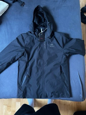 Svart vindjacka med huva från Arc'teryx - Snygg svart vindjacka från Arc'teryx med huva och dragkedja framtill. Jackan har två sidofickor med dragkedja och är tillverkad i ett vatten- och vindtätt material. Perfekt för blåsiga dagar och outdoor-äventyr.