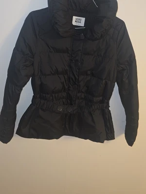 Svart pufferjacka från Vero Moda - Snygg svart pufferjacka från Vero Moda med hög krage och markerad midja med knappar. Jackan har quiltad design och är perfekt för kalla dagar. Stängs med dragkedja och tryckknappar framtill. Passar dig som vill ha en stilren och varm vinterjacka.