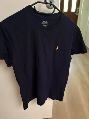 Marinblå t-shirt från Polo Ralph Lauren - Snygg marinblå t-shirt från Polo Ralph Lauren med klassisk gul broderad logga på bröstet. Tillverkad i mjuk bomull och har en normal passform med korta ärmar. Perfekt till jeans eller shorts för en clean och stilren look.
