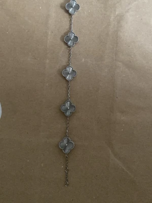 Silverfärgat armband med fyrklöver - Snyggt armband i silverfärg med fem fyrklöverformade länkar. Varje länk har en pärlliknande kant och en blank, metallisk yta i mitten. Kedjan är tunn och smidig, perfekt för att ge en elegant touch till din stil.