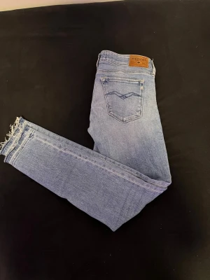Ljusblå jeans med rå kant - Säljer ett par ljusblå skinny jeans med klassisk femficksdesign och rå, fransad kant vid bensluten. Jeansen har en enkel bakficka med dekorativ söm och är tillverkade i mjukt denimtyg som ger en bekväm passform. Perfekta för en avslappnad och trendig look.