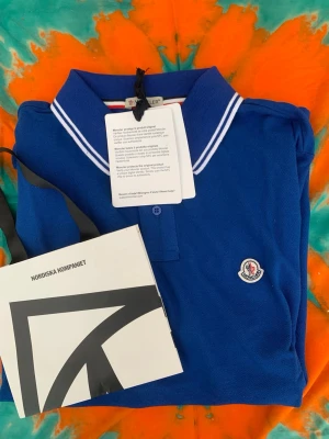 Blå pikétröja från Moncler - Snygg blå pikétröja från Moncler med vitrandig krage och Moncler-logga på bröstet. Klassisk polokrage med knappar framtill. Perfekt för dig som vill ha en stilren och sportig look.