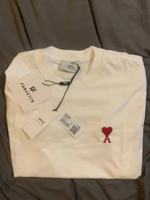 Vit t-shirt från Ami med rött hjärta - Vit t-shirt från Ami med klassisk passform och rund hals. På bröstet finns Ami:s ikoniska röda hjärta med bokstaven A broderat. Tillverkad i mjuk bomull och perfekt för en clean och stilren look.