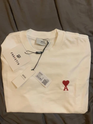 Vit t-shirt från Ami med rött hjärta - Vit t-shirt från Ami med klassisk passform och rund hals. På bröstet finns en liten röd broderad detalj med hjärta och bokstaven A. Tillverkad i mjuk bomull och perfekt för en clean och stilren look.