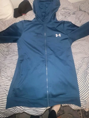 Blå hoodie från Under Armour - Snygg blå hoodie från Under Armour med dragkedja framtill och vit logga på bröstet. Tillverkad i ett mjukt och sportigt material som känns skönt mot huden. Utrustad med huva och två sidofickor. Perfekt för dig som gillar en clean och sportig stil.