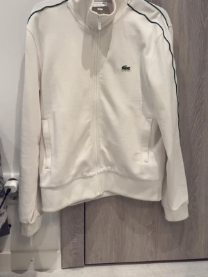 Vit zip-hoodie från Lacoste - Säljer en vit hoodie från Lacoste med hel dragkedja och gröna detaljer längs armarna. Använd några gånger. Man ser knappt att den är använd, bara om man kollar noggrant från jätte nära. Köpte den för 1900.  