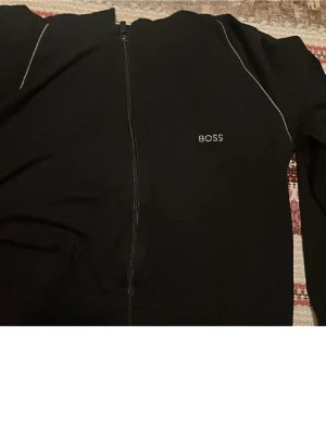 Svart hoodie med dragkedja från BOSS - Snygg svart hoodie från BOSS med dragkedja framtill och vit logga på bröstet. Hoodien har tunna vita detaljer längs axlarna. Perfekt för en chill och stilren look.