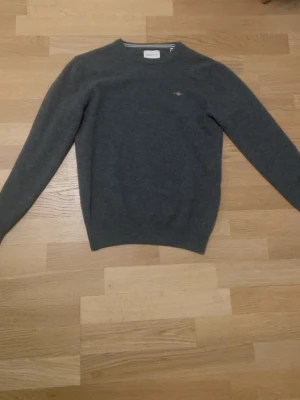 Mörkgrå stickad tröja från Gant - Mörkgrå stickad tröja från Gant med rund halsringning och diskret broderad logga på bröstet. Tröjan har ribbade muddar vid ärmslut och nederkant samt en klassisk passform. Perfekt för lager-på-lager och enkel att matcha.