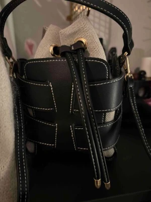 Svart och beige bucket bag med detaljer - Trendig bucket bag i svart läder med vita kontrastsömmar och beige tyginsats. Väskan har justerbar axelrem och snygga guldfärgade metalldetaljer. Perfekt för dig som vill ha en unik och stilren accessoar till din outfit.