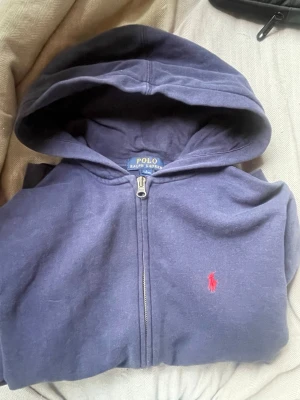 Mörkblå hoodie från Polo Ralph Lauren - Säljer en mörkblå hoodie med dragkedja från Polo Ralph Lauren. Klassisk design med huva, kängurufickor och den ikoniska röda loggan broderad på bröstet. Perfekt för en avslappnad och stilren look. storlek 14-16år