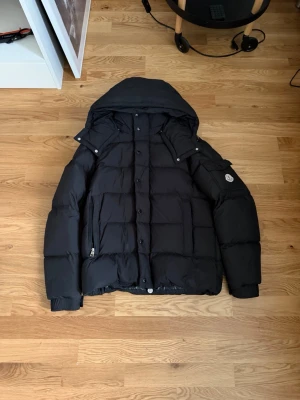Svart dunjacka från Moncler - Säljer en svart dunjacka från Moncler med avtagbar huva och stora fickor. Jackan har en quiltad design, tryckknappar och dragkedja framtill samt Moncler-logga på ärmen. Perfekt för kalla vinterdagar och riktigt snygg streetstil.