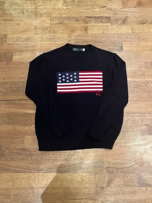 Mörkblå stickad tröja med USA-flagga - Mörkblå stickad tröja med rund hals och ribbade muddar. På bröstet finns en stor USA-flagga i rött, vitt och blått samt initialerna RL broderade i rött. Tröjan har en klassisk passform och är perfekt för dig som gillar stilrena plagg med en twist. Den står i storlek L men skulle säga mer M eller S