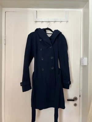 Mörkblå trenchcoat/kappa med huva - Stilren mörkblå trenchcoat med dubbelknäppning, huva och knytskärp i midjan. 