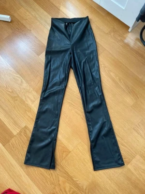 Svarta utsvängda byxor i fuskskinn - Säljer ett par svarta byxor i fuskskinn med hög midja och utsvängda ben. Snyggt glansigt material som ger en cool look. Perfekta för dig som vill sticka ut lite extra och gillar trendiga detaljer. Dom har också en liten slitt nere vid fötterna.                                                                                              Är öppen för att förhandla priset🌸