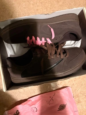 Nike bruna sneakers med rosa detaljer - Säljer ett par bruna Nike sneakers med svarta och rosa swoosh-loggor. Skorna har bruna och rosa skosnören samt perforerade detaljer på tån. Klassisk låg modell i mockaliknande material. Levereras i originalkartong.