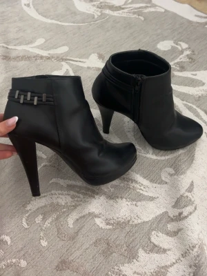 Svarta stilettklackade boots - Säljer ett par svarta stövletter med hög stilettklack och dragkedja på sidan. Snygg detalj med dekorativa band och metallspännen vid ankeln. Tillverkade i ett slätt material som ser ut som skinn. Perfekta för dig som vill ha en edgy och trendig look.