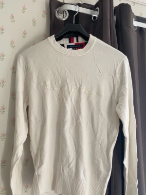 Vit stickad tröja Tommy Hilfiger - Snygg vit stickad tröja från Tommy Hilfiger med diskret broderad logga framtill. Tröjan har rund halsringning, långa ärmar och ribbade muddar vid ärmslut och nederkant. Perfekt för en clean och stilren look.