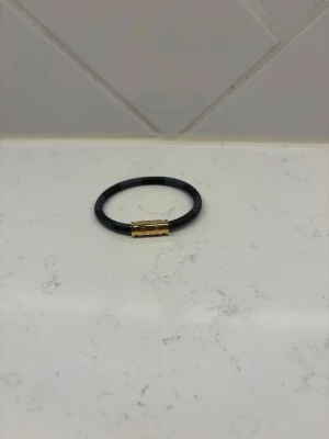 Svart armband med guldfärgat spänne - Stilrent svart armband i flätat läder med ett snyggt magnetlås i guldfärgad metall. Perfekt accessoar för att lyfta din outfit med en enkel men cool detalj. Passar både till jeans och skjorta. Armbandet är 17 cm långt. Hör av er gällande frågor!!