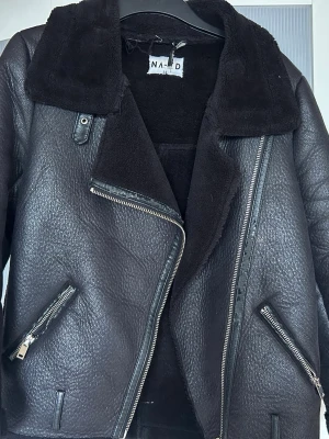Svart pälsjacka från NA-KD - Säljer en svart pälsjacka från NA-KD med bred krage och dragkedja framtill. Jackan har två snedställda fickor med dragkedjor och mjukt foder. Materialet är konstläder med fuskpäls på insidan och detaljer vid ärmslut och krage.