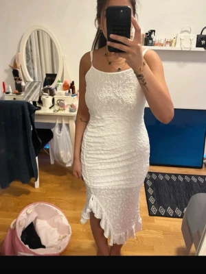 Vit långklänning med volangkant - Säljer en elegant vit långklänning med smala axelband och figurnära passform. Klänningen har en snygg volangkant nertill och ett diskret strukturmönster i tyget. Perfekt för dig som vill ha en stilren och trendig look.