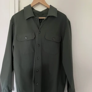 Olivgrön overshirt från Matinique - Snygg olivgrön overshirt från Matinique med klassisk krage och två bröstfickor med lock och knappar. Skjortan har långa ärmar och stängs med knappar framtill. Perfekt att bära som lager över en t-shirt.