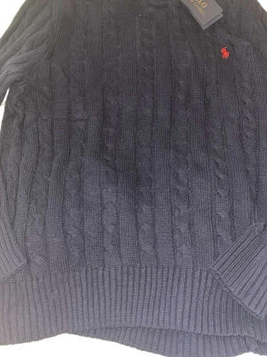 Mörkblå kabelstickad tröja Polo Ralph Lauren - Mörkblå kabelstickad tröja från Polo Ralph Lauren med klassisk rund halsringning och ribbade muddar. Tröjan har ett rött broderat logotyp på bröstet och är tillverkad i mjukt stickat material. Perfekt för att skapa en stilren look.