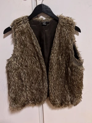 Brun fluffig pälsväst - Snygg brun pälsväst med fluffig fuskpäls och öppen front. Västen har en naturlig look med kort ludd och är perfekt för att ge outfiten extra edge. Passar till många olika stilar och är lätt att matcha med andra plagg.