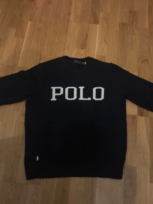 Mörkblå stickad Polo Ralph Lauren tröja - Svart stickad tröja med stor vit POLO-text på bröstet och liten broderad logga vid nederkanten. Klassisk rund halsringning och långa ärmar. Perfekt för dig som gillar stilrena och tidlösa plagg. Ralph lauren storlek L