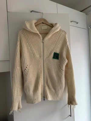 Beige/ vit stickad hoodie med dragkedja - Stickad beige hoodie med dragkedja framtill och ribbade muddar. Tröjan har en grön patch på bröstet och detaljerade stickningar. Långa ärmar med slitna kanter ger en avslappnad look. Zadig Voltaire. Nypris ca 3500+ kronor