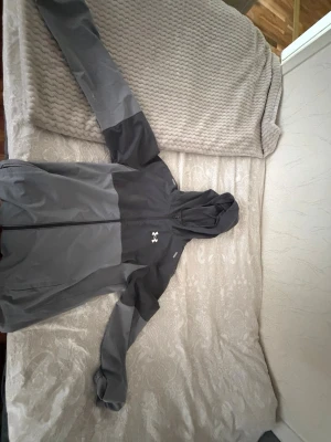 Tvåtonad grå vindjacka Under Armour - Vindjacka från Under Armour i två nyanser av grått med hel dragkedja och huva. Jackan har en sportig look, logga på bröstet och är gjord i lätt, vindtätt material. Perfekt för utomhusaktiviteter och streetwear. Köptes i JD för 1000kr
