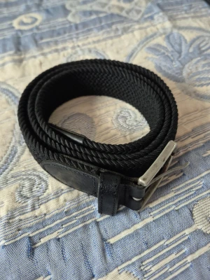 Dressmann bälte  - Snyggt svart flätat bälte från Dressmann med elastisk väv och detaljer i skinn. Perfekt för att ge din outfit en clean och stilren touch. Spännet är i silverfärgad metall och bältet är flexibelt för extra komfort.