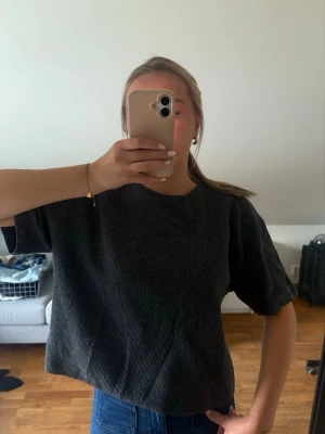 Mörkgrå ribbad croppad topp - En mörkgrå croppad topp med ribbad struktur och korta ärmar. Toppen har en loose passform och rund halsringning. Perfekt att styla med jeans eller kjol för en chill look.