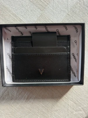 Svart kortfodral i läder från valentino - Snyggt svart kortfodral i läder med minimalistisk design och ett V i metall på framsidan. Fodralet har flera fack för kort och en smart pengaklämma i metall. Perfekt för dig som vill ha ett stilrent och smidigt sätt att bära dina kort.