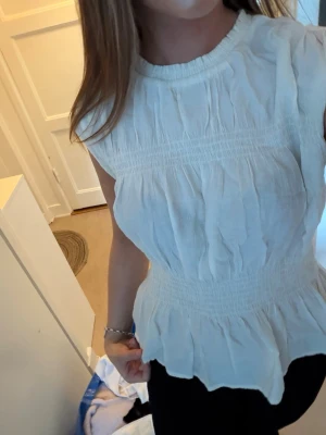 Vit blus med smock och volang - Säljer en söt vit blus med smockdetaljer i midjan och runt halsen. Blusen är ärmlös och har en volangkant nedtill som ger en feminin touch. Perfekt att styla med jeans eller kjol för en fräsch look.