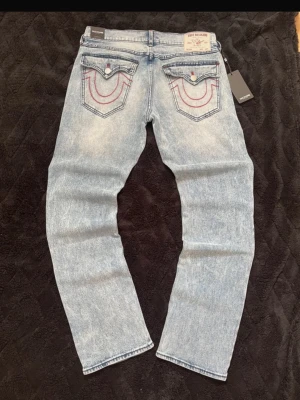 True religion jeans  - Säljer ett par ljusblå jeans med rak passform och tydliga kontrastsömmar i rött på bakfickorna. Jeansen har en klassisk femficksdesign och är tillverkade i denim med tvättad effekt för en cool vintagekänsla.