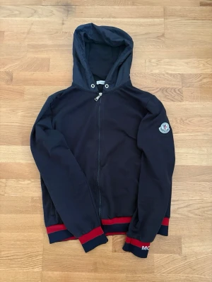 Marinblå tröja - Snygg marinblå hoodie med dragkedja och huva. Röd och marinblå rand. Moncler-logga på ärmen och diskret textdetalj vid mudden. Perfekt för dig som vill ha en stilren men ändå unik hoodie.