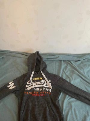 Mörkgrå Superdry hoodie med tryck - Snygg mörkgrå hoodie från Superdry med vit logga och text på bröstet samt färgglada detaljer. Huva med vita snören och tryck på ena ärmen. Perfekt för en avslappnad streetwear-look.