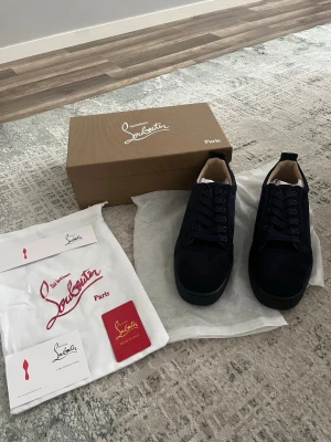 Mörkblå sneakers från Louboutin - Snygga mörkblå sneakers från Christian Louboutin i mocka med klassisk låg profil och svarta snören. Skorna har en stilren design och diskreta detaljer, samt den ikoniska röda sulan som märket är känt för. Perfekta för dig som vill ha något exklusivt och trendigt.