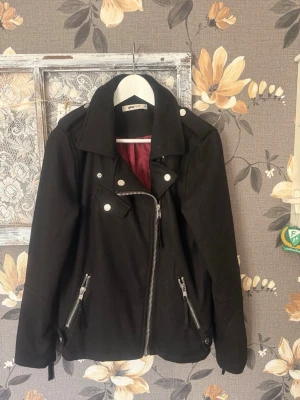 Svart bikerjacka med dragkedjor - Snygg svart bikerjacka med silvriga dragkedjor och detaljer. Jackan har bred krage, snedställd dragkedja framtill och två fickor med dragkedja. Perfekt för en cool och stilren look. Jackan är marinblå/svart. Jag upplever den mer svart dock. En snygg och varm höstjacka som även funkar vintertid